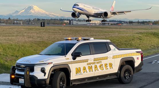 White pickup truck marked ‘Airport Duty Manager’ sits beside a runway with its roof lights on. In the background, an airplane is descending toward the runway, and a snow-covered mountain rises behind the landscape.