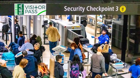 Travelers move through Security Checkpoint 3 at SEA Airport, placing items in bins while security staff operate screening equipment.