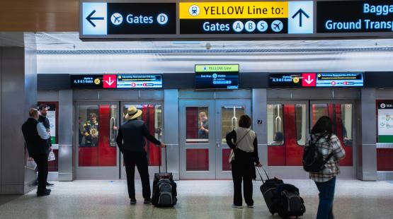 Passengers with rolling luggage stand on a polished floor facing closed doors of an airport underground shuttle train. Overhead signs display directions, including arrows toward Gates D and Gates A, B, S, and a yellow line indicator. The train’s interior and a person inside are visible through the glass doors.