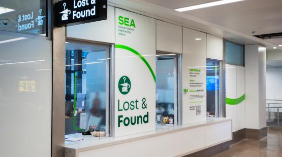 A Lost and Found service area at SEA Airport, with a bright overhead sign reading “Lost & Found” and a large wall display featuring the SEA airport logo and contact information. The space includes service windows, counters with small items, and glass partitions reflecting light from the terminal.