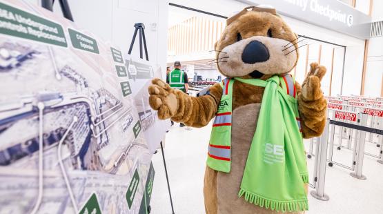 The SEA Airport mascot JETT, wearing a reflective safety vest and a bright green scarf with “SEA Seattle–Tacoma International Airport” printed on it, stands inside an airport security area. JETT gestures toward a large construction project display board set up on an easel. Behind JETT, airport staff in reflective vests are visible near signage for “Security Checkpoint 6” and barriers labeled “Emergency Exit Only.