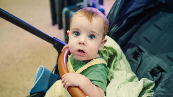 A small child sits in a stroller indoors, holding onto a brown handlebar and covered with a light green blanket. Luggage and other travel items are visible in the background.
