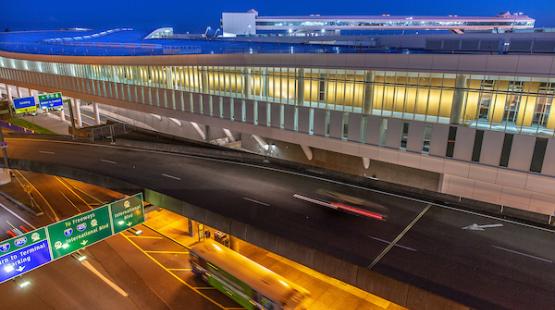 Elevated airport roadway at night with blurred headlights and taillights from moving vehicles beneath a long, modern terminal building with illuminated windows. Overhead signs mark directions to arrivals, the terminal, and parking.
