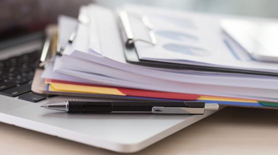 A stack of documents on clipboards rests on top of an open laptop, with a black pen placed across the laptop’s surface.