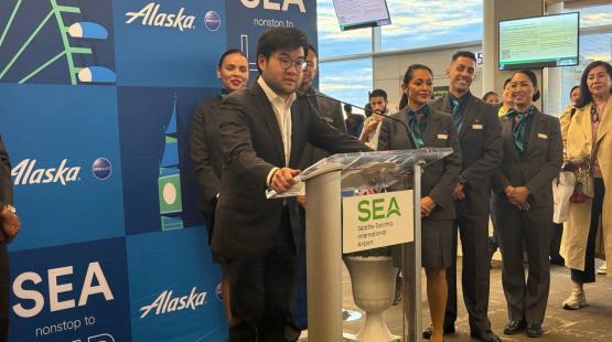 Port of Seattle Commissioner Sam Cho speaks at a podium in front of a banner celebrating Alaska Airlines' inaugural flight to London.