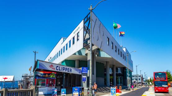 Side angle view of the Port of Seattle’s Headquarters Building at Pier 69, showing the FRS Clipper entrance, street signage, recycling bins, and a red double‑decker tour bus parked along the roadway.