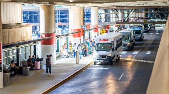 Airport curbside pickup area with shuttle vans lined up, passengers waiting with luggage, and signs marking different pickup zones under a covered roadway.
