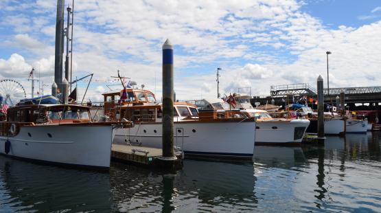 A row of classic wooden motorboats is docked at a marina on calm water, with tall mooring posts in front and a partly cloudy sky above. Colorful signal flags hang from several boats, and a large Ferris wheel and pier structures are visible in the background.