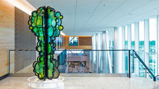 Artist Fumi Amano’s sculpture “Shared Breath” is shown on Level 3 of the C Concourse Expansion. The artwork consists of layered, circular green and yellow glass-like forms arranged vertically on a white base. It is displayed in a bright interior space with wood paneling, large windows, and views of seating and structural elements below.